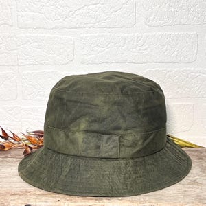 May include: An olive green bucket hat with a textured, waxed-style finish. The hat features a flat crown, a wide brim, and a matching fabric band around the base of the crown. The material displays subtle variations in colour and a creased appearance.