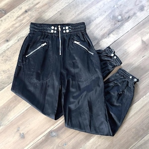 May include: Black satin trousers with a zip and silver button accents. The trousers have a drawstring waist and side pockets.