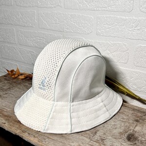 May include: A white bucket hat featuring a combination of solid fabric panels and breathable mesh sections. The hat includes a light blue embroidered kangaroo logo on the front panel and is constructed with stitched seams along the brim.