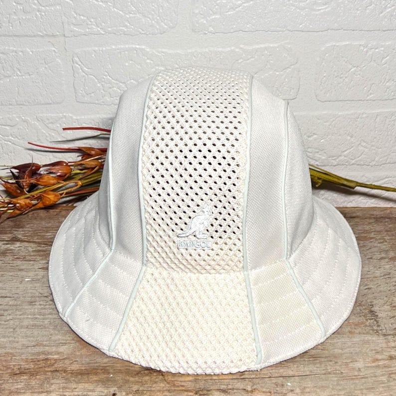 May include: A white bucket hat featuring a central vertical mesh panel and solid side panels. The front of the hat displays an embroidered Kangol kangaroo logo in white thread. The design includes a wide brim and structured seams throughout the crown.