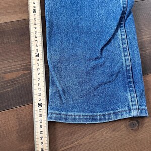 May include: Close-up of blue denim fabric with a straight hem. A wooden ruler with inch markings from 58 to 88 is positioned next to the denim. The fabric is on a dark brown wooden surface.