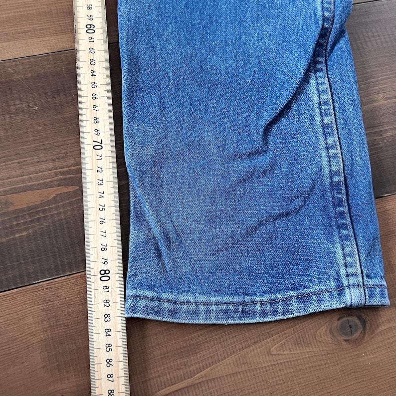 May include: Close-up of blue denim fabric with a straight hem. A wooden ruler with inch markings from 58 to 88 is positioned next to the denim. The fabric is on a dark brown wooden surface.