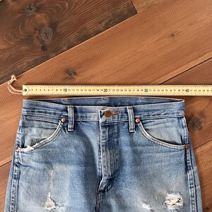 May include: Close-up of light blue denim jeans with distressed details, including rips and fading. The jeans have a classic five-pocket design with copper-colored rivets and a button closure. A wooden ruler is placed across the top of the jeans.