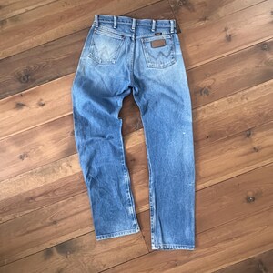 May include: Light blue denim jeans with a classic five-pocket design. The jeans feature a brown leather patch on the back pocket and a faded wash. The jeans are laid flat on a wooden surface.
