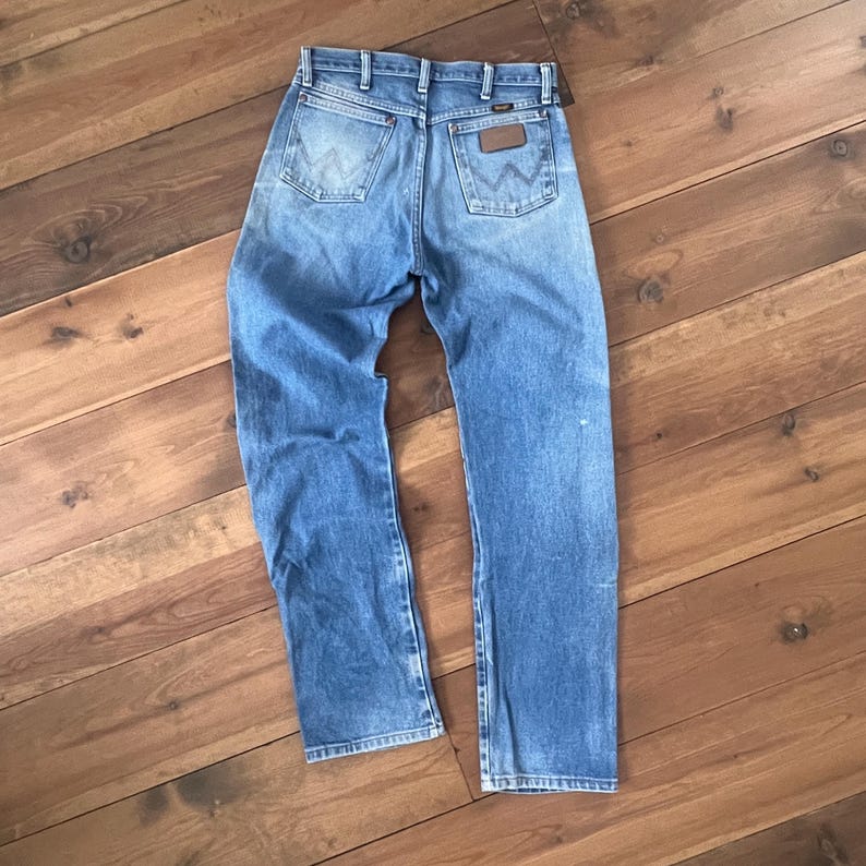 May include: Light blue denim jeans with a classic five-pocket design. The jeans feature a brown leather patch on the back pocket and a faded wash. The jeans are laid flat on a wooden surface.