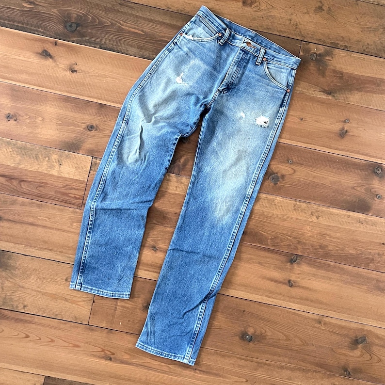 May include: Light blue denim jeans with distressed details, including faded areas and small rips. The jeans have a classic five-pocket design and a straight leg cut. The jeans are laid flat on a wooden surface.