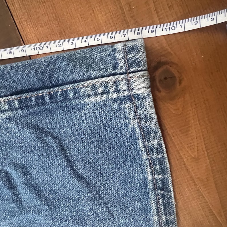 May include: Close-up of a section of blue denim jeans with a measuring tape laid across the fabric. The tape shows measurements in inches. The jeans are on a wooden surface.