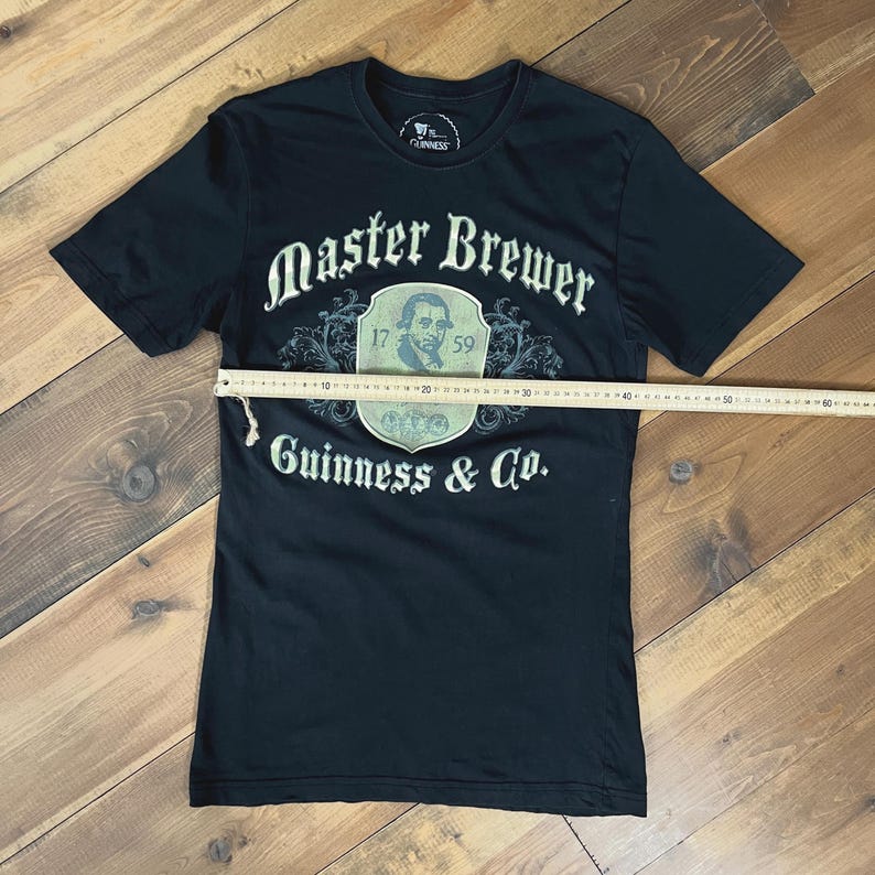 以下が含まれることがあります： 「Master Brewer」と「Guinness & Co.」の文字が白でプリントされた黒いTシャツ。肖像画と数字「17」と「59」が入った紋章が特徴です。Tシャツは定規で測定されています。