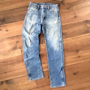 May include: Light blue denim jeans with a faded wash and distressed details. The jeans have a classic five-pocket design, a straight leg, and a button and zipper closure. The jeans are laid flat on a wooden surface.