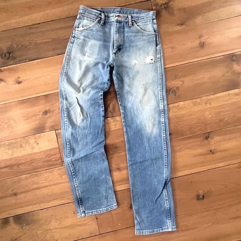 May include: Light blue denim jeans with a faded wash and distressed details. The jeans have a classic five-pocket design, a straight leg, and a button and zipper closure. The jeans are laid flat on a wooden surface.