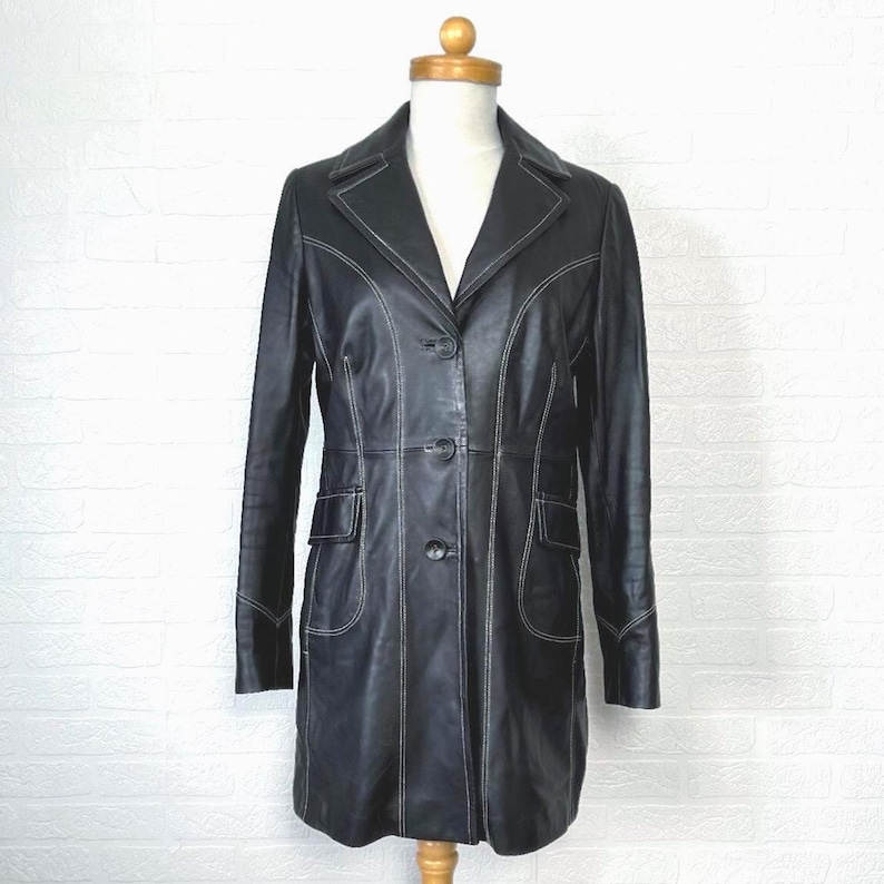 May include: A black leather coat with white stitching. The coat has a long, fitted silhouette and a collar. It features a button closure and two pockets.