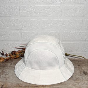 May include: A white bucket hat featuring a combination of smooth fabric panels and a textured mesh crown. The hat has a wide, downward-sloping brim and a structured design, showcasing a clean, monochromatic aesthetic with visible stitching details.