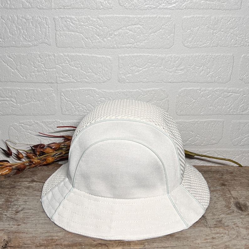 May include: A white bucket hat featuring a combination of smooth fabric panels and a textured mesh crown. The hat has a wide, downward-sloping brim and a structured design, showcasing a clean, monochromatic aesthetic with visible stitching details.