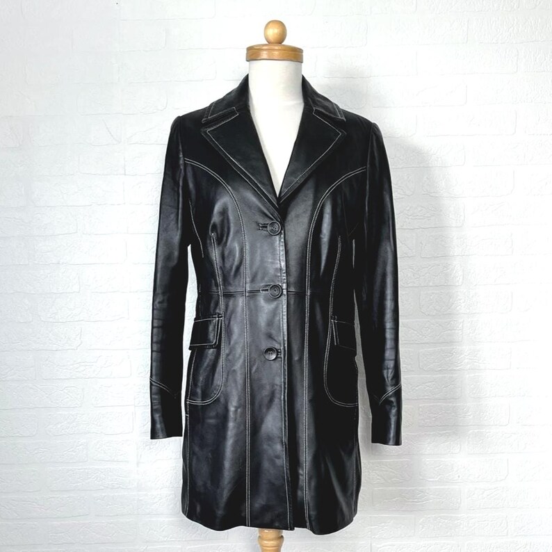 May include: A black leather coat with white stitching. The coat has a long, fitted silhouette and a button-up front.