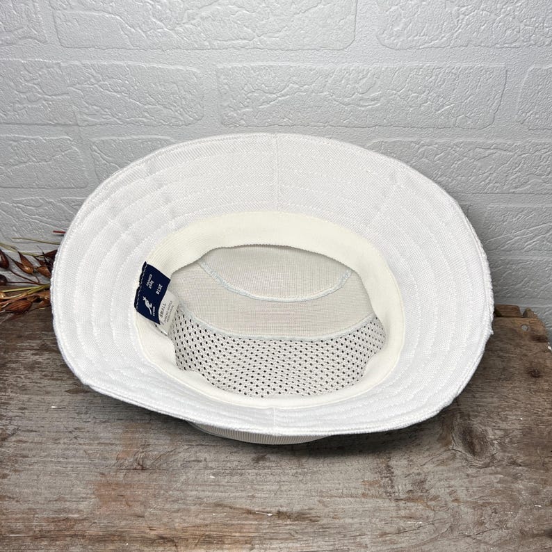 May include: A white bucket hat viewed from the inside, featuring a textured fabric exterior, a white interior sweatband, and a mesh lining at the crown. A small dark blue tag with a white kangaroo logo and the text "KANGOL" is attached to the band.