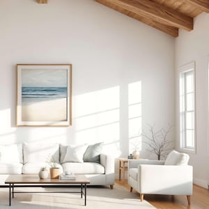 May include: A bright, airy living room with white walls, a white sofa, and a framed beach scene artwork. A wooden coffee table and a white armchair complete the space. The room has a natural, minimalist aesthetic.