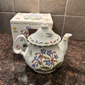 May include: A white porcelain teapot with a floral design featuring blue, pink, and yellow flowers. The teapot has a lid and a spout. The teapot is sitting on a table.