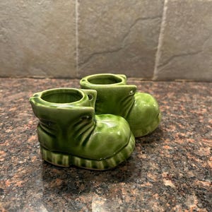May include: Two small green ceramic boots with a textured surface. The boots are shaped like children's shoes and have a rounded toe.