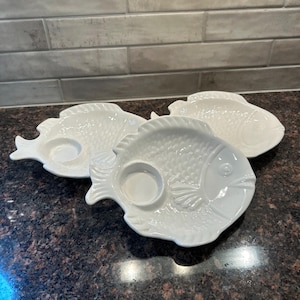 May include: Three white ceramic fish-shaped serving dishes. Each dish has a circular indentation, possibly for holding a dip or sauce. The dishes are arranged on a dark countertop, suggesting a kitchen or dining setting. The fish design adds a nautical theme.