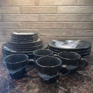 May include: A complete black ceramic dinnerware set with a textured finish. The set includes plates, bowls, and mugs. The mugs have handles and a white wave design. The plates and bowls are stacked neatly.