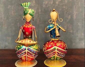 May include: Two colourful metal figurines of people playing musical instruments. The figurine on the left is playing a drum, and the figurine on the right is playing a flute. Both figurines are decorated with intricate patterns and bright colours.