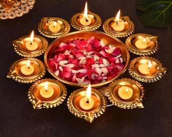 May include: A gold metal diya set with nine individual candle holders arranged in a circular pattern. The diya is filled with pink and white flower petals.