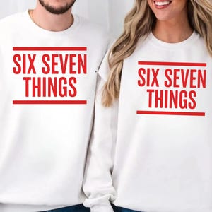 May include: White crew neck sweatshirts featuring the red text "SIX SEVEN THINGS". The text is framed by red lines. The sweatshirts are a matching set, suitable for couples or friends.