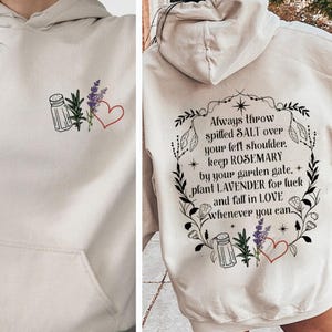May include: A beige hooded sweatshirt featuring a small graphic on the front and a large graphic on the back. The back design includes a floral wreath with a salt shaker, rosemary, lavender, a red heart, and the text: Always throw spilled SALT over your left shoulder, keep ROSEMARY by your garden gate, plant LAVENDER for luck and fall in LOVE whenever you can.