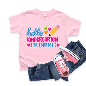 May include: A pink t-shirt with the words "hello KINDERGARTEN I'M (NAME)" in blue and pink, with a pencil and heart graphics. The shirt is paired with blue jeans and pink sneakers.