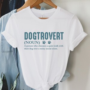 May include: White t-shirt with a blue graphic that says "Dogtrovert (NOUN) A person who chooses a quiet walk with their dog over a noisy social event."
