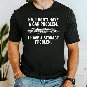May include: A black short-sleeve t-shirt featuring white text that reads: NO, I DON'T HAVE A CAR PROBLEM. I HAVE A STORAGE PROBLEM. Between the two lines of text is a white graphic illustration of four different car models lined up side by side.