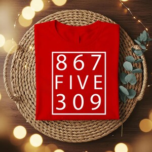 May include: A bright red t-shirt featuring a white rectangular border containing the white text 867 FIVE 309 arranged in three lines. The shirt has a crew neckline and short sleeves, presented in a flat lay style with a clean, bold graphic design.