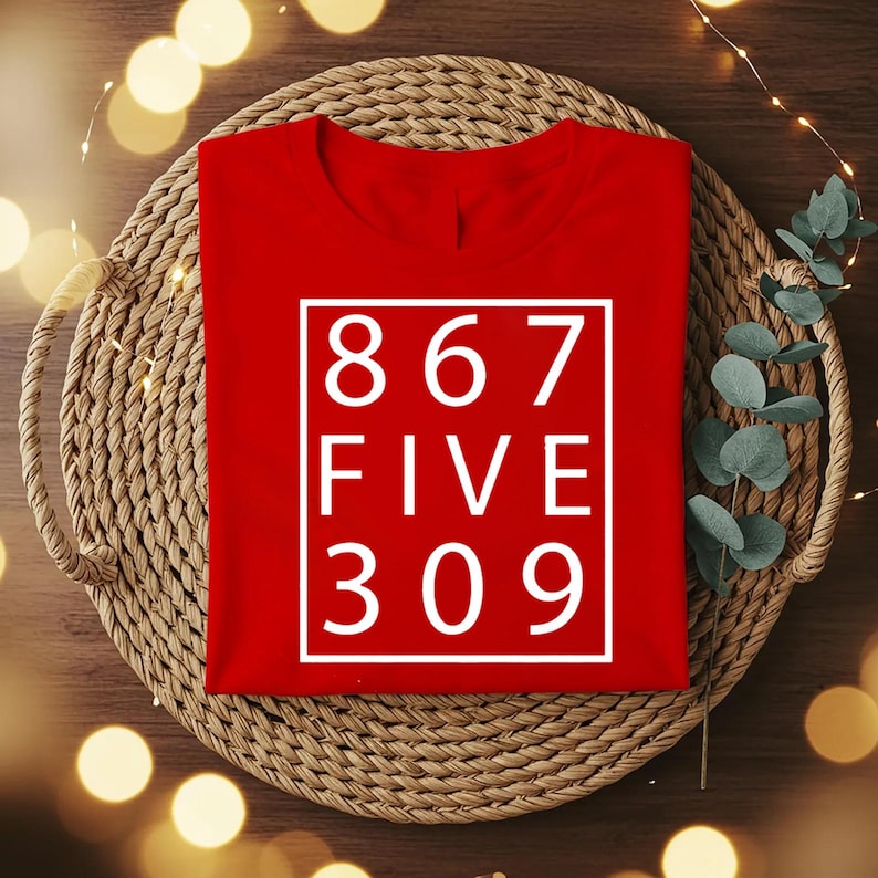 May include: A bright red t-shirt featuring a white rectangular border containing the white text 867 FIVE 309 arranged in three lines. The shirt has a crew neckline and short sleeves, presented in a flat lay style with a clean, bold graphic design.
