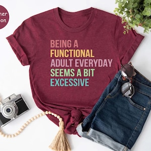 Puede incluir: Una camiseta color granate con el texto "Ser un adulto funcional todos los días parece un poco excesivo" en colores arcoíris.