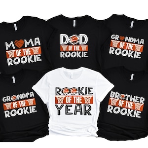 May include: Six black and white t-shirts with basketball-themed text. The shirts read "Mama of the Rookie", "Dad of the Rookie", "Grandma of the Rookie", "Grandpa of the Rookie", "Rookie of the Year", and "Brother of the Rookie".