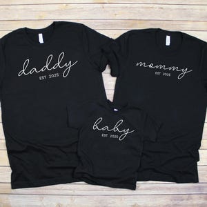 Puede incluir: Tres camisetas negras con texto blanco en cursiva: "daddy", "mommy" y "baby", cada una con "EST 2025" debajo. Las camisetas están juntas, sugiriendo un conjunto familiar.