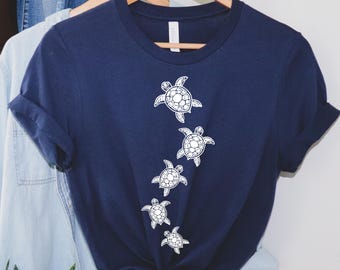 May include: Navy blue t-shirt featuring a design of five white sea turtles in a vertical pattern. The shirt is hanging on a wooden hanger. The shirt is made of a soft material and has rolled-up sleeves.
