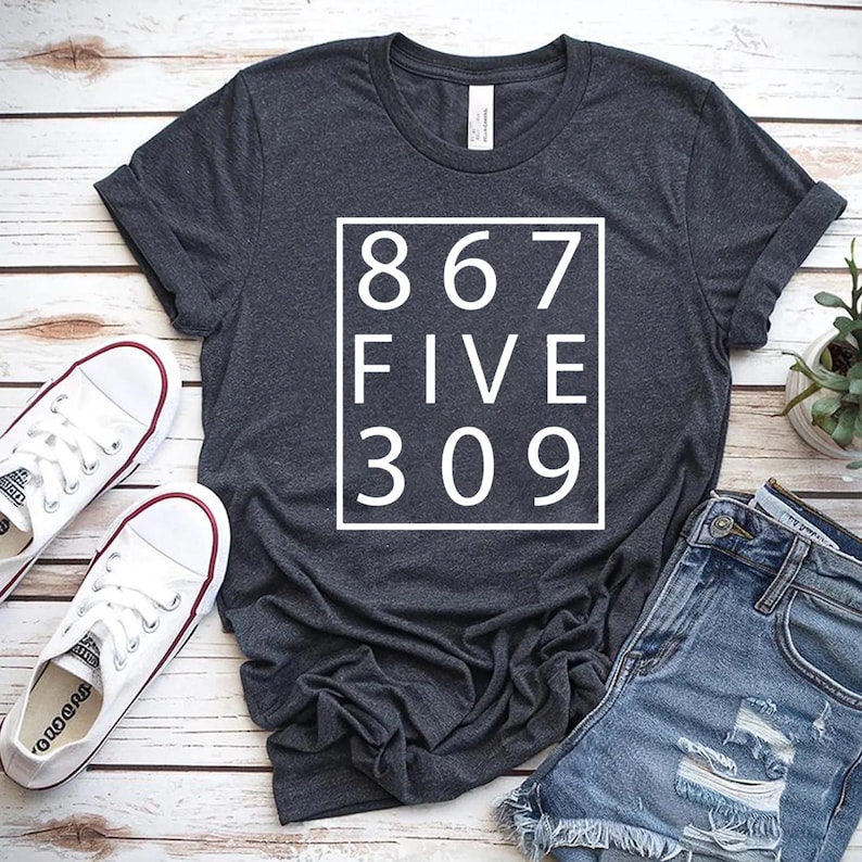 May include: A dark heather gray short-sleeve t-shirt featuring a white square outline containing the text 867 FIVE 309 in a clean sans-serif font. The shirt is displayed flat with a pair of white canvas sneakers and blue denim shorts.