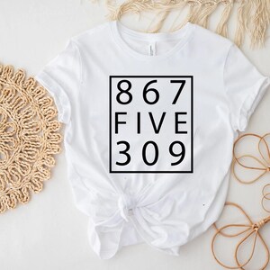 May include: A white short-sleeved t-shirt featuring a black square graphic on the chest. Inside the square, the text reads 867, FIVE, and 309 stacked in three rows. The shirt has a crew neckline and is tied in a knot at the bottom front hem.
