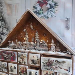 May include: A wooden advent calendar shaped like a house with a peaked roof. The top features a miniature train, pine trees, and a crescent moon. The front has numbered drawers decorated with festive illustrations, including plaid patterns, winter scenes, and holly berries.