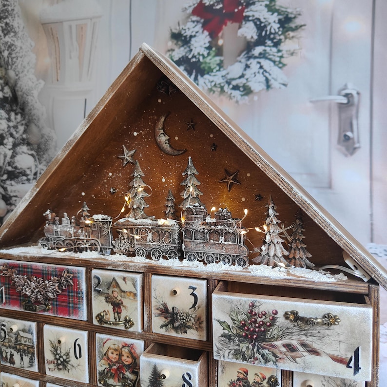 May include: A wooden advent calendar shaped like a house with a peaked roof. The top features a miniature train, pine trees, and a crescent moon. The front has numbered drawers decorated with festive illustrations, including plaid patterns, winter scenes, and holly berries.