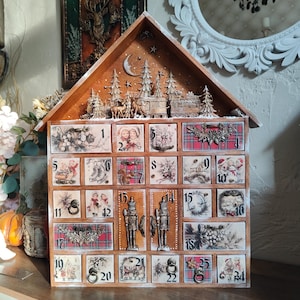 May include: A wooden house-shaped advent calendar with twenty-four numbered drawers. The top features a carved winter scene with trees, deer, and a train. Each drawer displays vintage-style holiday illustrations, plaid patterns, and metal pulls.