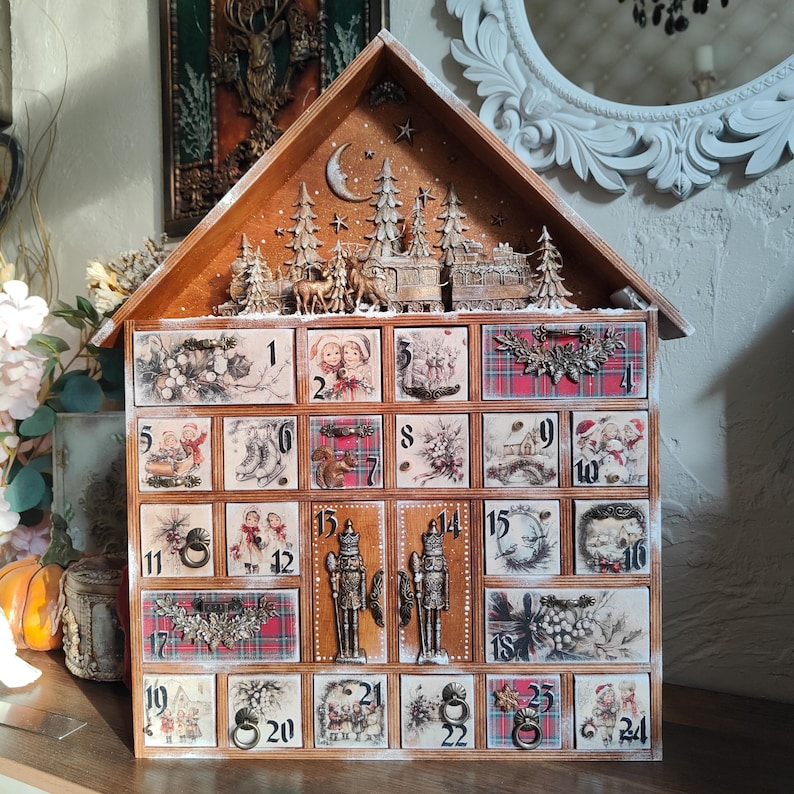 May include: A wooden house-shaped advent calendar with twenty-four numbered drawers. The top features a carved winter scene with trees, deer, and a train. Each drawer displays vintage-style holiday illustrations, plaid patterns, and metal pulls.