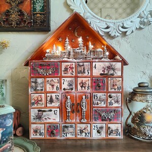 May include: A wooden house-shaped advent calendar with twenty-four numbered drawers featuring vintage-style holiday illustrations, plaid patterns, and metal pulls. The top section displays a miniature train and snowy trees illuminated by warm string lights.