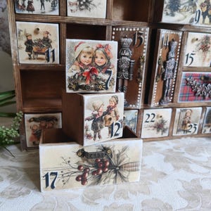 May include: A wooden advent calendar with numbered drawers featuring vintage-style illustrations of children, nutcrackers, and holly berries. The drawers are stacked in front of the main unit, displaying the numbers 7, 12, and 17 with ornate metal handles.