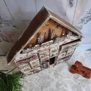 May include: A wooden house-shaped advent calendar with a peaked roof, featuring twenty-four small drawers numbered one through twenty-four. The front displays festive illustrations, a miniature train, and integrated warm LED string lights.