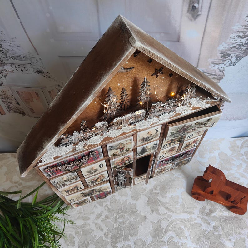 May include: A wooden house-shaped advent calendar with a peaked roof, featuring twenty-four small drawers numbered one through twenty-four. The front displays festive illustrations, a miniature train, and integrated warm LED string lights.