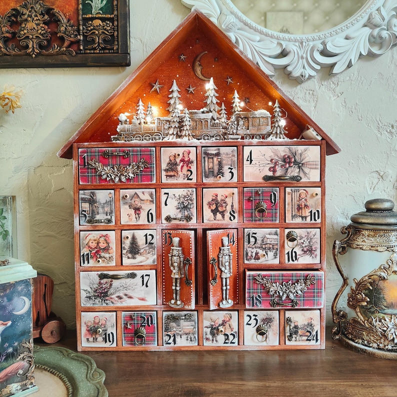 May include: A wooden house-shaped advent calendar with twenty-four numbered drawers. The front features vintage-style winter illustrations, red plaid patterns, and metal hardware. A miniature train and forest scene with warm LED lights sits on the roof.