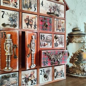 May include: A wooden advent calendar with twenty-four numbered drawers featuring various holiday illustrations, including nutcrackers, winter scenes, and plaid patterns. Each drawer has a metal handle or pull, and the unit is finished in a warm brown tone.