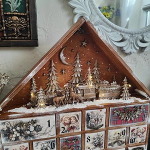 May include: A wooden advent calendar shaped like a house with a triangular top featuring a relief of a train, deer, and pine trees under a crescent moon and stars. The base has numbered drawers decorated with vintage-style holiday illustrations.